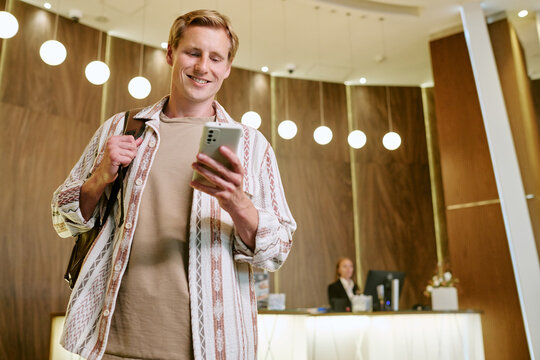 Smiling young adult Caucasian man using smartphone in hotel lobby, checking in online. Low angle shot representing modern travel technology and digital hospitality services