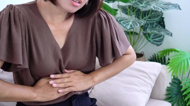 Asian woman touching her stomach feeling severe stomach ache, abdominal pain, cramps, or indigestion symptoms while sitting on couch at home. Healthcare, illness, and medical problem concept