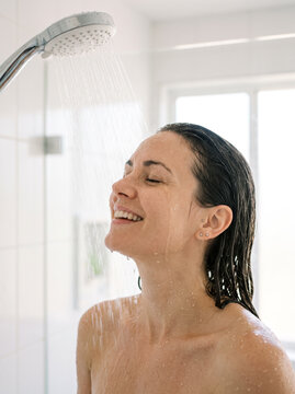 an adult woman happily taking a shower, only her head, shoulders and upper neck visible, leaning forward toward a rainfall shower stream, smiling with eyes closed, wet hair and realistic water droplet