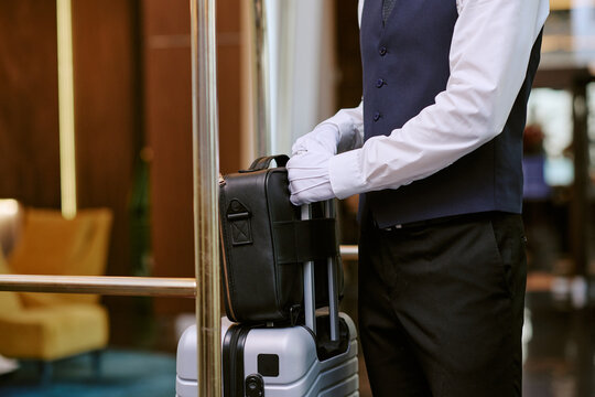 Cropped shot of male hotel bellhop wearing gloves while placing black leather bag on luggage cart. Image represents luxury hospitality service and premium guest experience