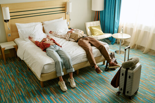 Young adult Caucasian couple lying on bed in hotel room after arrival, high angle view. Image represents travel fatigue and vacation comfort, ideal for hospitality marketing