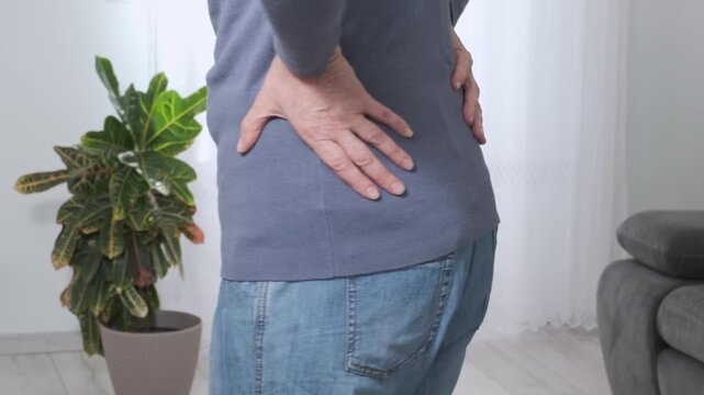 Back pain. Middle aged woman holding her lower back and massaging it with her hands at home. Close-up, rear view, side view. Lumbago, sciatica, lower back pain, lumbago in the back, hernia, protrusion