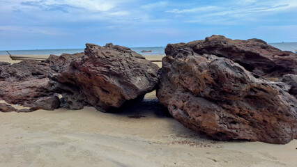 Close perspective of two heavy porous rock boulders sitting on soft beach sand with a distant wooden fishing boat on the calm ocean horizon. © Bendy Moe