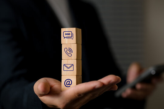 Close-up of a person holding a smartphone in one hand while balancing vertically stacked wooden blocks with communication icons representing messaging, calling, email, and online interaction.