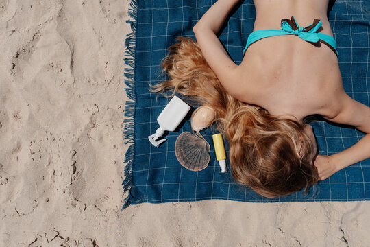 Blonde woman lies face down on a blue beach towel beside two hair protection products and shells on the sand. Sun and salt water hair care on holiday.