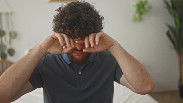 Man rubbing his eyes and nose with hands while sitting on bed in bedroom beside plant and nightstand; fatigue.