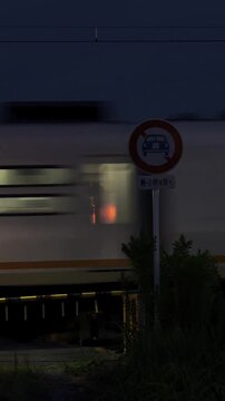 Express train speeding through a Japanese railway crossing at dusk with flashing warning signal lights and road sign