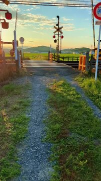 Gravel path leading to a Japanese rural railway crossing with warning signals and road sign against scenic countryside sunset