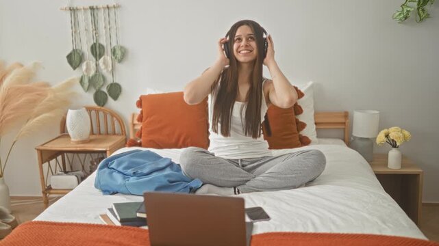 Woman smiling and adjusting headphones while cross legged on bed with laptop and backpack in bedroom; relaxed study focus.