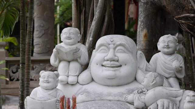 Laughing budai statue surrounded by small child figures in a tropical garden with palms and trees. Symbol of abundance.