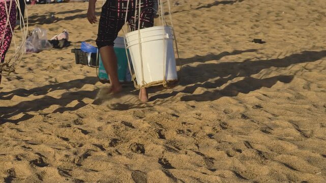 Authentic rural coastal life at sunrise with side lighting, featuring a local vendor carrying heavy seafood buckets on a shoulder pole along the sandy beach in Phu Yen, Vietnam