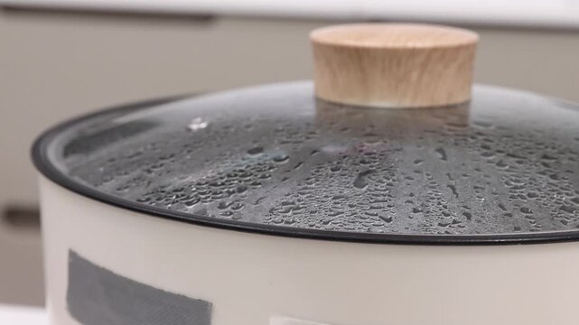 Close-up shot of an electric skillet or hot pot lid with boiling water inside, heavy condensation droplets on the glass, and steam escaping from the rim.