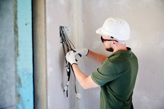 Electrician connecting wires inside wall junction box and organizing cables. Worker in helmet and gloves preparing electrical system for installation, interior construction and wiring setup concept