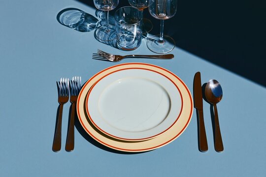 Formal place setting with stacked plates and silverware on a cool grey tabletop, featuring crisp shadows and red rim accents for dining catalogs