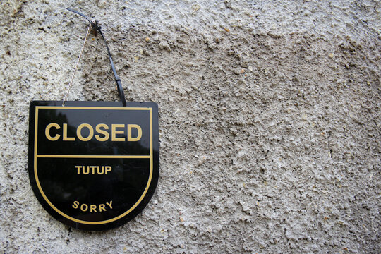 a bilingual hanging sign in Bahasa Indonesia and English indicating that a business is temporarily closed, mounted on a textured concrete wall.