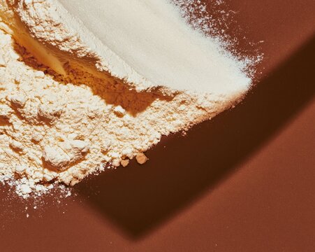 Loose flour and icing sugar drifts on a deep cocoa brown surface, featuring a powdery texture and hard shadows for food packaging and backgrounds