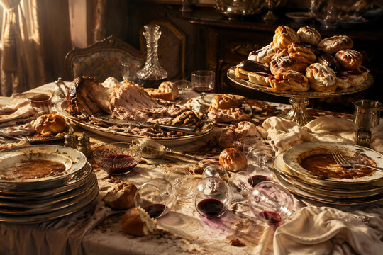 Wide still-life of overloaded banquet table after long meal, torn roast, stacked plates, scattered bread, tipped wine glasses and pastries, gluttony mood
