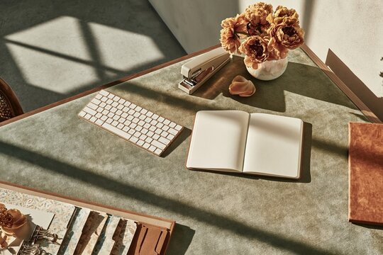 Open blank notebook on a muted blue desk with a keyboard, peonies, and stationery, glowing in natural sunlight for a mockup and home office branding