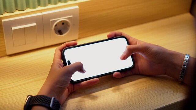 Hand holds phone with white screen. Phone rests on wooden surface near wall outlet. Warm light illuminates the scene. User taps screen with finger. Natural lighting adds cozy atmosphere