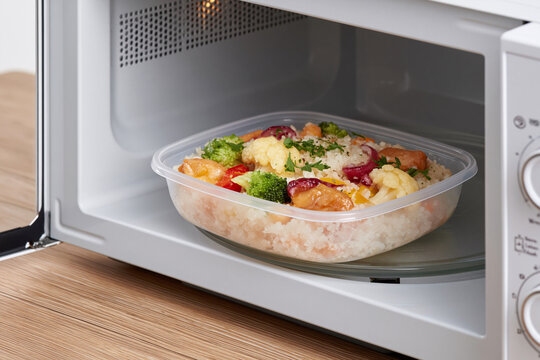 Microwave oven holds a colorful meal. Plastic dish contains rice, vegetables, and protein. Fresh ingredients look appetizing and ready to heat. No people present, so emotions are irrelevant