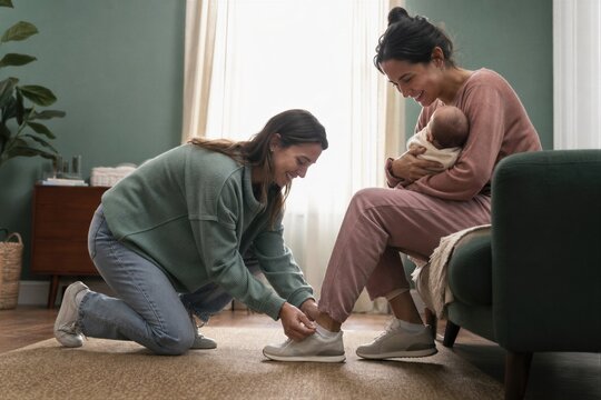 Woman friend helping new mother holding her newborn baby by tying shoelace on sneaker. Postpartum care and support for family at home. Concept of maternal love and friendship.