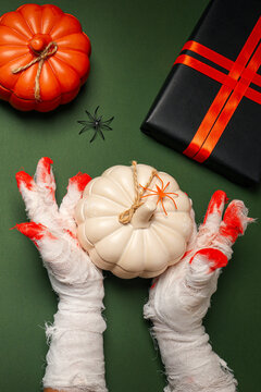 Halloween. Mummy hands grasp white pumpkin while surrounded by spooky decor