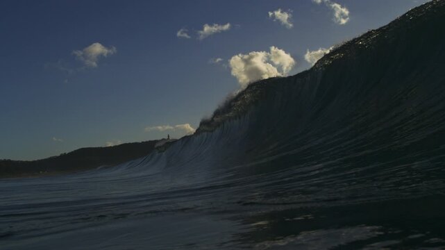 Dark ocean wave rising against blue sky and sunlit clouds in super slow motion