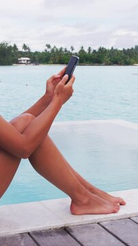 Phone lifestyle. Woman using phone relaxing by pool on beach in on vacation. Lady using phone app at luxury travel holidays resort. Raw vertical video of real woman. unrecognizable woman.