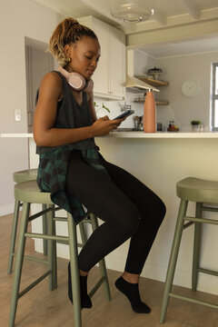 African woman sitting on green stool in kitchen using smartphone, wearing pink headset