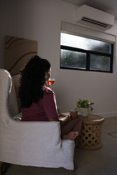 Indian woman sitting in maroon loungewear on upholstered armchair holding glass teacup by window