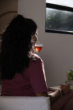 African American woman sitting on cushioned chair at home, holding glass teacup, notebook on lap