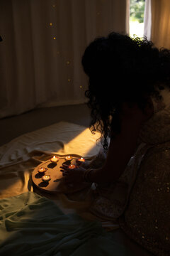 Indian woman in embellished dress arranging lit diyas on wooden tray by window, copy space