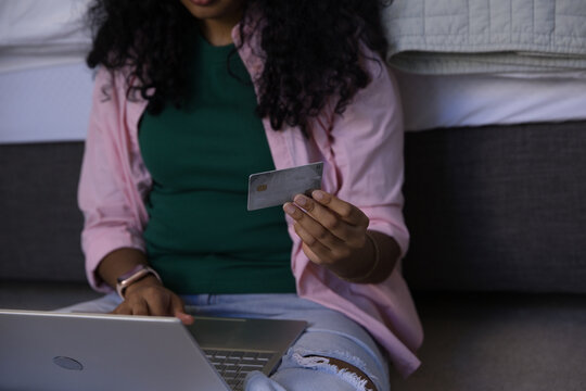 Female sitting on floor at foot of bed wearing ripped jeans holding bank card using laptop