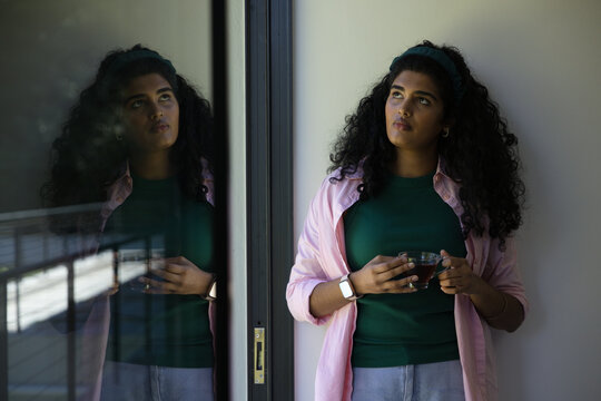 Indian woman standing at home beside sliding glass door holding clear cup and gazing, copy space