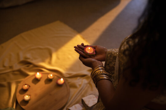 Indian woman holding lit diya in hands on floor with embroidered sleeve and bangles, copy space