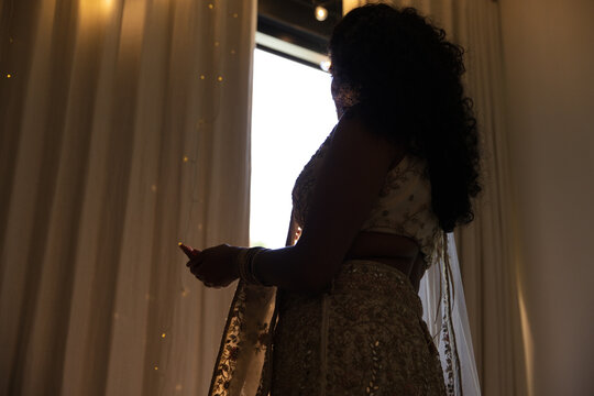 Indian woman standing by curtained window holding smartphone in sari and bangles, copy space