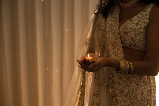 Indian woman holding lit diya with both hands against curtain backdrop, lehenga, bangles