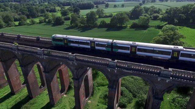 Close aerial drone view of a passenger train crossing a historic brick railway viaduct, surrounded by lush green countryside, trees and fields in beautiful natural daylight