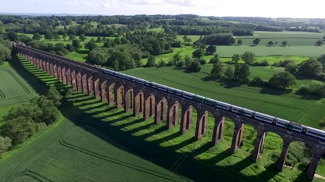 Aerial drone view of a passenger train crossing a historic brick railway viaduct through lush green countryside. Scenic rural landscape with rolling fields, trees and traditional railway architecture
