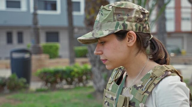 Woman soldier in army uniform talking on smartphone, profile face visible and hand holding phone to ear, camouflage cap and tactical vest on a street near buildings; duty determination.