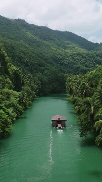 Vertical drone slowly ascends behind a Loboc River cruise boat traveling downstream between forested hills in Bohol