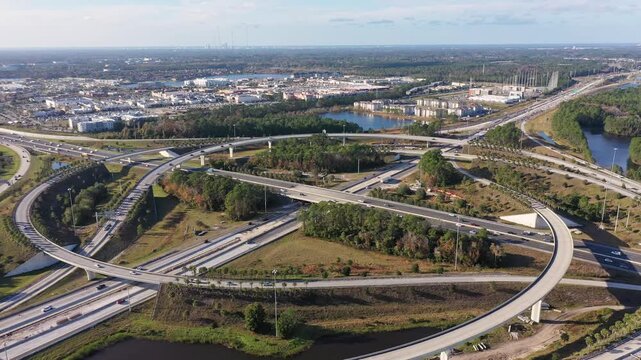 Expansive aerial footage overlooking a multi-level highway interchange as vehicles travel through sweeping curved ramps and intersecting freeways surrounded by waterways and urban development.