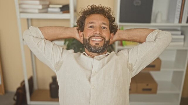 Man smiling with hands behind head in a studio office, leaning back in a casual shirt; work break relaxation.