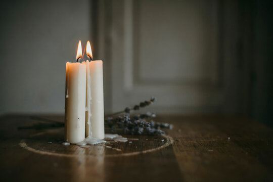 Two tall white pillar candles burning with bright flames on a dark wooden table with dried lavender sprigs in a dim, moody interior room setting
