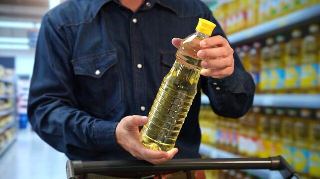 Man holding a bottle of cooking oil while shopping in a supermarket aisle with a cart, comparing grocery products on store shelves.