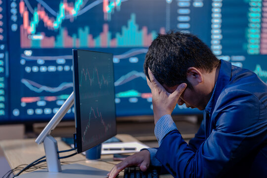 Stressed financial trader holds his head at a desk with stock charts on multiple screens, showing market loss, investment risk and business pressure.