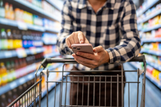 Man using a smartphone while pushing a shopping cart through a supermarket aisle, comparing products or managing a grocery list.