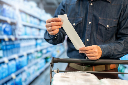 Man in a denim shirt checks a shopping receipt beside a cart in a supermarket aisle, comparing household products and prices.