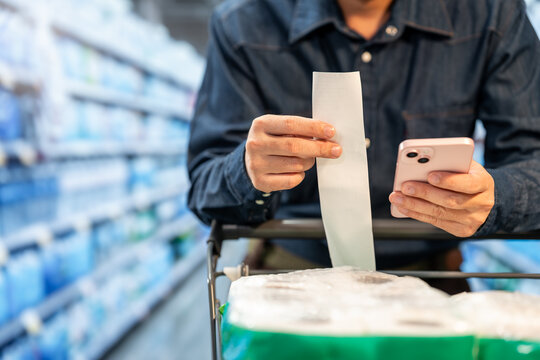 Close-up of a shopper in a supermarket checking a long receipt on a smartphone beside a cart with groceries, showing budgeting and purchase review.