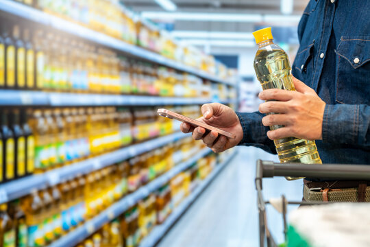Man comparing cooking oil prices on a smartphone in a supermarket aisle, holding a bottle beside a shopping cart.